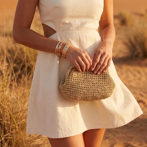May include: A cream-colored sleeveless dress with side cutouts paired with a gold-toned woven clutch bag and a stack of thin gold-toned metal bangles worn on the wrist. The clutch features a structured frame closure and a textured, braided finish.