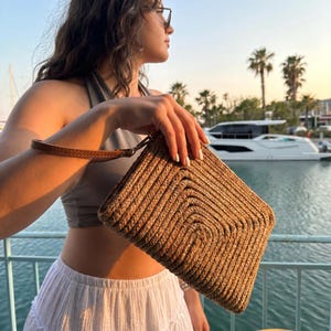 May include: A woven straw clutch with a brown leather wrist strap. The clutch has a square shape with a textured, braided design. The background shows a waterfront scene with palm trees and boats.