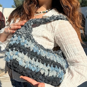 May include: A denim shoulder bag in shades of blue and black. The bag is made from woven strips of denim, creating a textured, patchwork design. The strap is also made of denim. The bag is held by a person.