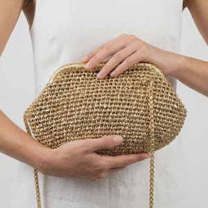 May include: A gold-colored crocheted clutch bag with a metallic sheen, featuring a structured frame closure and a thin, matching gold-tone chain shoulder strap. The bag has a textured, woven surface and a rounded, semi-circular silhouette.
