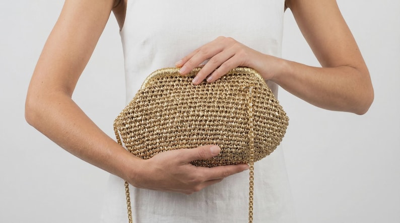 May include: A gold-colored crocheted clutch bag with a metallic sheen, featuring a structured frame closure and a thin, matching gold-tone chain shoulder strap. The bag has a textured, woven surface and a rounded, semi-circular silhouette.
