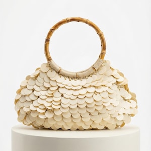 Puede incluir: Bolso de mano color crema con asa de bambú. El bolso está cubierto de pequeñas lentejuelas redondas superpuestas de color crema. El asa es de color marrón claro. El bolso está sobre un pedestal blanco.