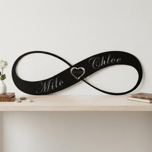 May include: A black metal infinity symbol wall sign featuring the engraved names Milo and Chloe in a cursive script. A central heart shape contains the engraved year 2027. The sign is a continuous loop design with a smooth matte finish.
