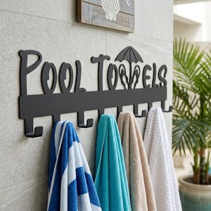 May include: A black metal wall-mounted rack with the text Pool Towels, featuring a small umbrella and flip-flop icon. The rack has seven hooks, with four hanging towels in blue and white stripes, solid turquoise, solid tan, and solid white.