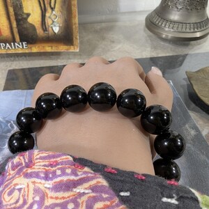 Puede incluir: Un brazalete de cuentas negras con cuentas grandes y redondas. El brazalete se lleva en la muñeca, mostrando la superficie brillante y pulida de las cuentas. Las cuentas son de tamaño uniforme y están ensartadas.