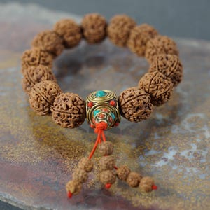 May include: A beaded bracelet featuring brown, textured beads. The bracelet has a decorative central bead with turquoise and red accents, and a red tassel with smaller beads. The bracelet is displayed on a mottled brown and gray surface.