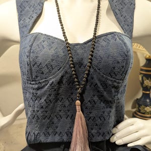 Op de afbeelding: Een donkerblauwe bustier top met een geometrisch patroon wordt op een mannequin getoond. Een lange kralenketting met een kwast hangt over de top. De kwast heeft een lichtroze kleur.