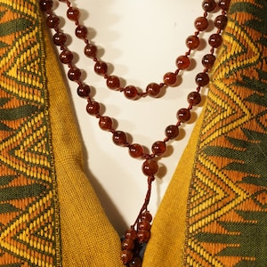 May include: A double strand of brown gemstone beads hangs on a person's chest. The beads are strung on a brown cord. The person is wearing a green and gold patterned garment.