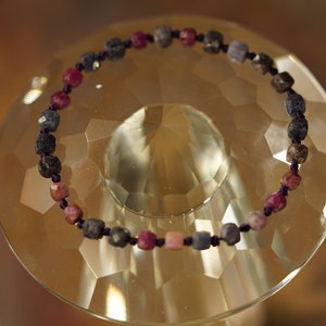 Op de afbeelding: Een armband gemaakt van kleine, vierkante kralen in tinten roze, paars en grijs. De kralen zijn geregen aan een donkerpaarse koord.