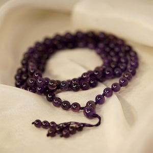 May include: A purple amethyst mala bead necklace with a tassel. The necklace is made of round beads and is lying on a white fabric background.