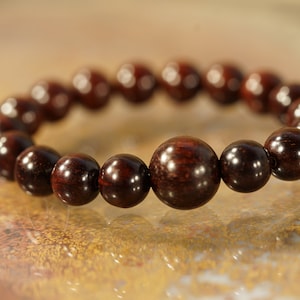 May include: A dark brown beaded bracelet. The bracelet is made of polished, round wooden beads. The beads vary slightly in size, with a larger central bead. The bracelet is displayed on a light brown surface.