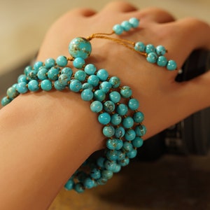 May include: A turquoise beaded bracelet with a tassel. The bracelet is made of multiple strands of beads and is tied with a gold cord.