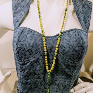 May include: A long beaded necklace with yellow and green beads hangs against a blue patterned top. The necklace features a tassel at the end. The top is a bustier style with a geometric design.