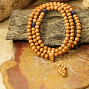 May include: A wooden mala prayer bead necklace with blue beads interspersed throughout. The necklace is displayed on a brown stone surface.