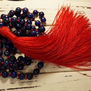 May include: A blue lapis lazuli mala bead necklace with a red tassel. The necklace is made of 108 beads and is strung on a red cord.