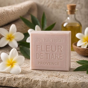 Könnte beinhalten: Ein hellrosa Seifenstück mit dem Text "100% Huile Végétale FLEUR DE TIARE PROVENCE". Die Seife ist von weißen Blumen mit gelben Zentren, grünen Blättern und einer Ölflasche umgeben. Die Seife liegt auf einer Steinoberfläche.