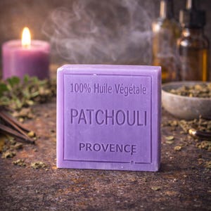 Op de afbeelding: Een lavendelkleurig Patchouli-zeepblok met de tekst "100% Huile Végétale" en "Patchouli Provence" in reliëf op de voorkant. Een brandende paarse kaars en andere aromatherapie-artikelen staan op de achtergrond.