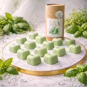 Peut inclure: Une douzaine de bonbons verts en forme de cœur disposés sur un plateau en marbre avec un bord doré. Un contenant cylindrique en carton avec les lettres "LBA" et une illustration botanique est derrière les bonbons. Des feuilles de basilic frais et des cristaux entourent le plateau.