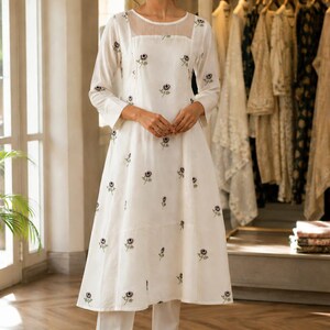 May include: A white knee-length tunic dress with long sleeves and a sheer mesh yoke, featuring small embroidered dark floral motifs scattered across the fabric. The dress is paired with matching white straight-leg trousers and light-coloured sandals.