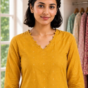 May include: A mustard yellow long-sleeved tunic featuring a V-neckline adorned with small, matching fabric pom-pom trim. The fabric has a subtle, repeating white geometric dot pattern throughout, with three-quarter length sleeves and a straight hem.