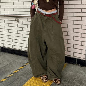 May include: Olive green wide-leg trousers with a red belt. The high-waisted trousers have a relaxed fit. The person is wearing a brown long-sleeved top and leopard print shoes. A small purse hangs from the shoulder.