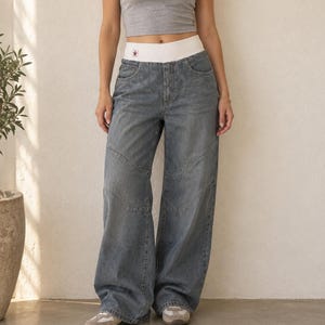 May include: Wide-leg denim jeans in a faded blue wash featuring a thick white elastic waistband with a small red star logo patch. The jeans have a high-rise fit, front pockets, and curved seam detailing across the thighs and knees.