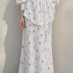 May include: A two-piece set consisting of a long-sleeved button-down shirt with a collar and a matching floor-length skirt. Both pieces are made of a white fabric featuring a repeating pattern of small pink flowers and thin green stems.