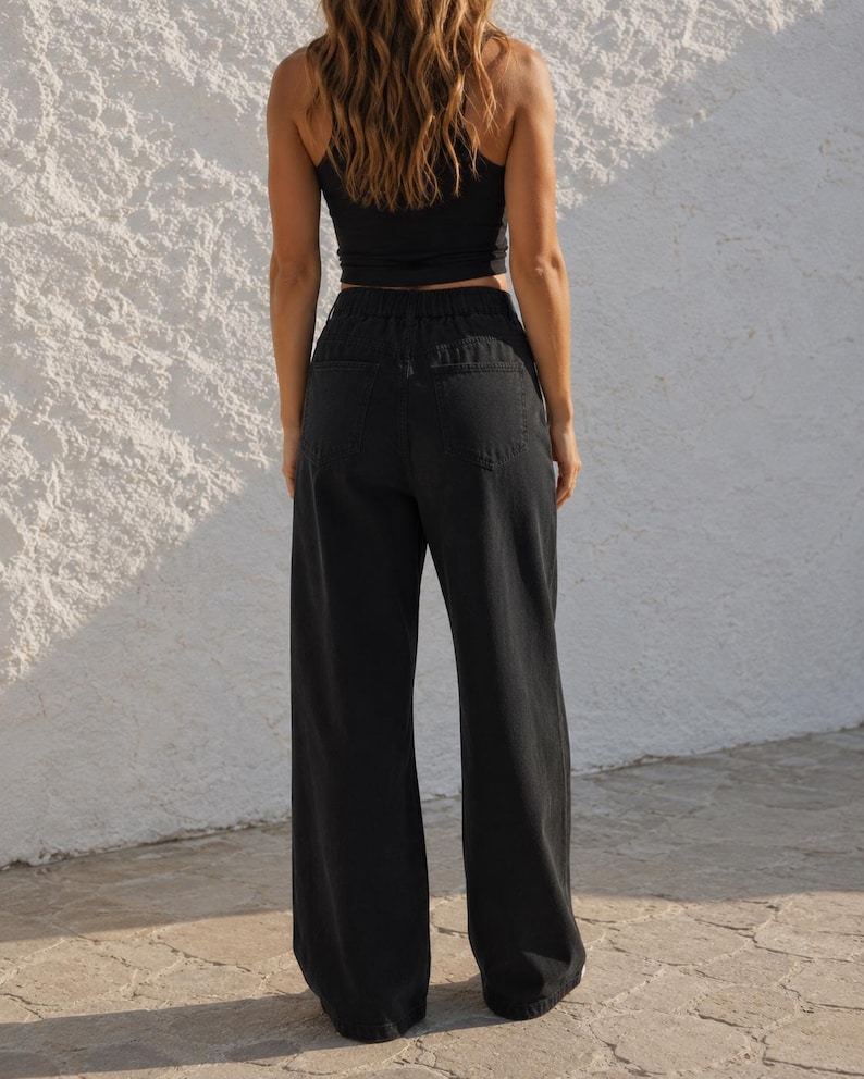 K&ouml;nnte beinhalten: Schwarze Jeans aus Denim mit hoher Taille, elastischem Bund, zwei aufgesetzten Ges&auml;&szlig;taschen und weit geschnittenen Beinen. Die Hose wird mit einem schwarzen &auml;rmellosen Oberteil kombiniert und zeigt eine schlichte, funktionale Gestaltung.