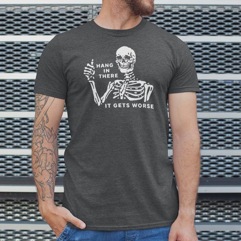 Pu&ograve; includere: T-shirt grigio scuro con uno scheletro bianco che alza il pollice. Il testo sulla maglietta dice "Hang in There" e "It Gets Worse".