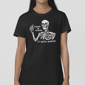 Pu&ograve; includere: T-shirt nera con uno scheletro bianco che fa il pollice in su. Il testo "HANG IN THERE" &egrave; sopra lo scheletro e "IT GETS WORSE" sotto. Un design umoristico.