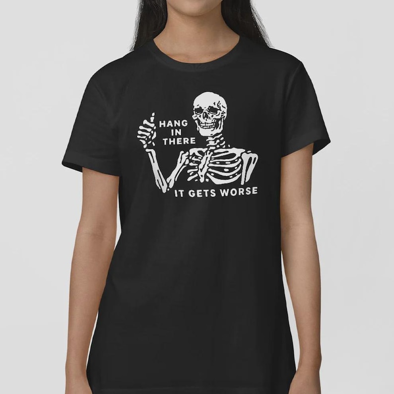 Pu&ograve; includere: T-shirt nera con uno scheletro bianco che fa il pollice in su. Il testo "HANG IN THERE" &egrave; sopra lo scheletro e "IT GETS WORSE" sotto. Un design umoristico.
