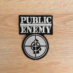 Puede incluir: Parche bordado en blanco y negro con las palabras "PUBLIC ENEMY" sobre un diseño de diana. El parche es rectangular con un emblema circular debajo, sobre un fondo de madera.