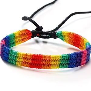 May include: A woven bracelet featuring a vibrant rainbow pattern with horizontal stripes of red, orange, yellow, green, blue, and purple. The bracelet includes a black adjustable cord closure with two thin black strings extending from the knot.