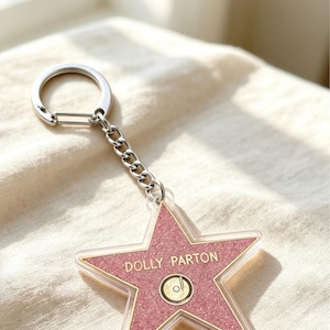 May include: A star-shaped keyring featuring a pink glittery surface with the name DOLLY PARTON printed in gold lettering above a gold circular icon. The charm is attached to a silver-toned metal chain and a circular split ring for keys.