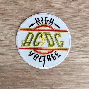 Peut inclure: Écusson rond blanc avec l'inscription "HIGH VOLTAGE" et "AC/DC" en lettres noires et dorées. L'écusson comporte une bande horizontale rouge et or au centre. Un éclair est placé entre les lettres "A" et "C".