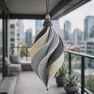 Puede incluir: Un adorno de ganchillo colgante con un diseño en espiral en forma de lágrima. La pieza está construida con paneles alternos de hilo gris oscuro, gris claro y color crema. Está suspendida por un cordón gris con una pequeña cuenta de madera arriba.