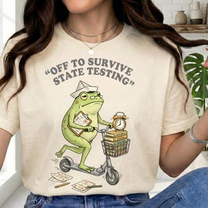 Puede incluir: Camiseta color crema con la frase gris "OFF TO SURVIVE STATE TESTING". El diseño presenta una rana verde montada en un patinete, con un sombrero de papel y llevando libros y una carpeta.