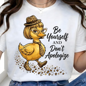 May include: White t-shirt featuring a cartoon duck wearing glasses, a hat, and a tie with a leopard print design. The text "Be Yourself and Don't Apologize" is written in black script.