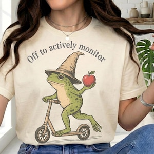 May include: Cream-colored t-shirt with the text "Off to actively monitor." The graphic features a green frog wearing a wizard hat, riding a scooter, and holding a red apple. The design has a whimsical, cartoonish style.