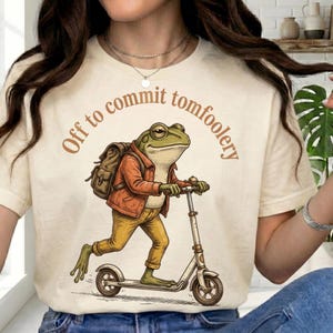 Op de afbeelding: Crèmekleurig T-shirt met de tekst "Off to commit tomfoolery" in bruin. De afbeelding toont een cartoonkikker op een scooter, met een jas en rugzak. De kikker is groen, met een gele broek en een oranje jas.