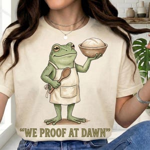 May include: A cream-colored t-shirt featuring a green frog wearing a beige apron, holding a wooden spoon and a basket of dough. Below the illustration, the text reads "WE PROOF AT DAWN" in bold, dark green capital letters.