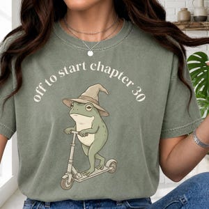 May include: Sage green t-shirt featuring a cartoon frog riding a scooter and wearing a wizard hat. The text "off to start chapter 30" is arched above the frog. The shirt is made of a soft material.