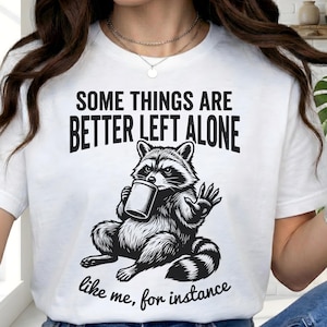 Puede incluir: Camiseta blanca con un gráfico negro de un mapache sosteniendo una taza y saludando. El texto en la camiseta dice "SOME THINGS ARE BETTER LEFT ALONE" y "like me, for instance."