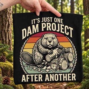 May include: A black t-shirt featuring a graphic of a beaver working on a pile of logs with a tripod and toolbox. The design includes a sunset background and the text "IT'S JUST ONE DAM PROJECT AFTER ANOTHER" in a bold, arched, light-colored font.