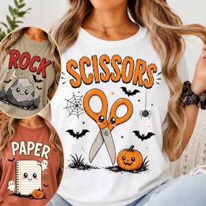 May include: A white t-shirt featuring the orange text SCISSORS above a cartoon pair of scissors with a face, a jack-o'-lantern, a spider web, and bats. Insets show a tan shirt with the text ROCK and a rust shirt with the text PAPER.