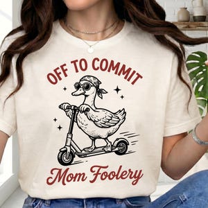 Puede incluir: Camiseta color crema con un gráfico de un pato en un scooter, con gafas de sol y un pañuelo. El texto "OFF TO COMMIT" y "Mom Foolery" está en color granate.