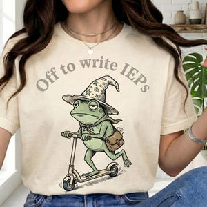 Könnte beinhalten: Cremefarbenes T-Shirt mit dem Text "Off to write IEPs". Das Shirt zeigt einen Cartoon-Frosch mit Zaubererhut, der Roller fährt. Der Frosch hat eine Umhängetasche und einen grünen Schal.
