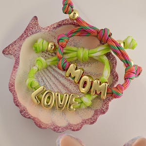 Pode incluir: Três pulseiras coloridas da amizade com letras douradas que soletram "LOVE" e "MOM" são exibidas em um prato decorativo em forma de concha. As pulseiras apresentam cordões verdes, rosa, laranja e brancos com detalhes dourados.