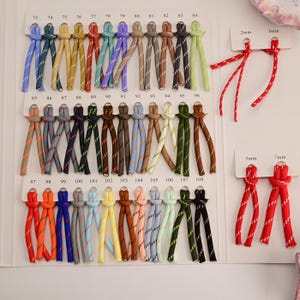 May include: A color chart featuring numerous pairs of braided cords in various shades, each labeled with numbers 73 through 108. To the right, four red cords are displayed on cards labeled 2mm, 3mm, 5mm, and 7mm, indicating their thickness.