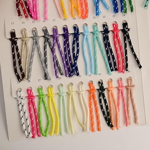 Op de afbeelding: Een kleurenstaalkaart met talloze paren gevlochten koorden in diverse kleuren, elk door genummerde sleuven gestoken. De koorden vertonen een consistent wit gespikkeld patroon op effen kleuren, waaronder rood, blauw, groen, geel en roze.